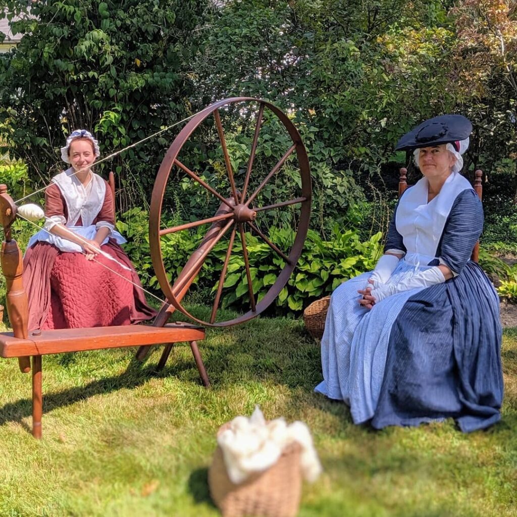 Two women, wearing historical outfits, sit next to a spinning wheel during a previous LexSeeHer event.